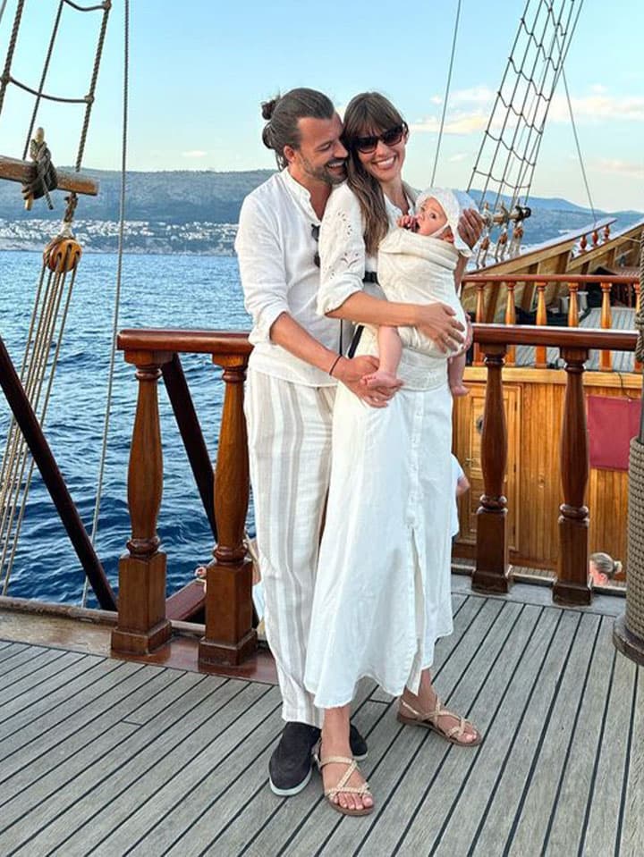 A couple in white outfits stands on a wooden ship deck, holding a baby. The ocean and distant shoreline are visible under a clear sky.