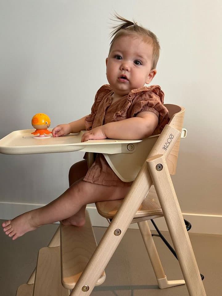 Baby sitting in a wooden KLAPP high chair, wearing a brown outfit, with a small toy on the tray.